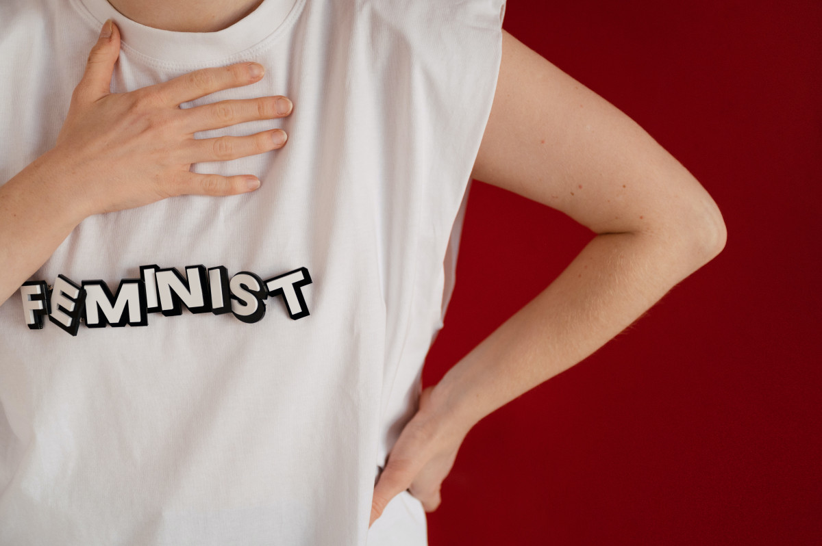 A white woman's torso. One arm cocked, the other hand on her chest. She wears a white tshirt with the word Feminist on it.